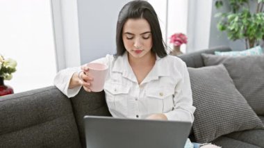 Genç bir İspanyol kadın rahat bir oturma odasında laptopuyla çalışırken bir fincan kahvenin keyfini çıkarıyor..