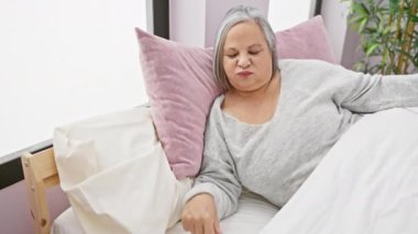 Yaşlı kadın uyandıktan sonra yatakta geriniyor, yatak odası arka planda bitkilerle dolu.