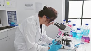 Olgun bir bilim kadını, parlak bir laboratuvar ortamında mikroskop altında bir numuneyi inceliyor..