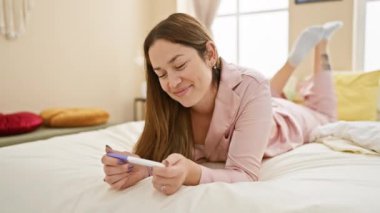 Gülümseyen bir kadın yatakta uzanıyor, hamilelik testini iyi aydınlatılmış, rahat bir yatak odasında bekliyor..