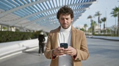 Sakallı yakışıklı İspanyol adam güneşli bir şehir parkında palmiye ağaçlarıyla akıllı telefon kullanırken gülümsüyor..