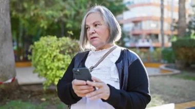 Yeşillik ve arka planda binalar olan bir parkta akıllı telefon kullanan olgun bir kadın.