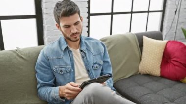 Rahat İspanyol bir adam gündelik giysiler içinde bir tablet tutuyor, modern bir oturma odasında bir kanepede oturuyor..