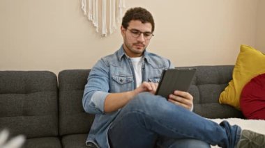 Gözlüklü ve sakallı genç bir adam, rahat bir şekilde kanepede otururken tablet kullanıyor..