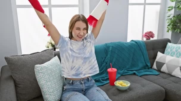 若いブルネットの女性は居心地の良いリビングルームでソファーに座っている間 興奮して赤いスカーフを振ります — ストック動画
