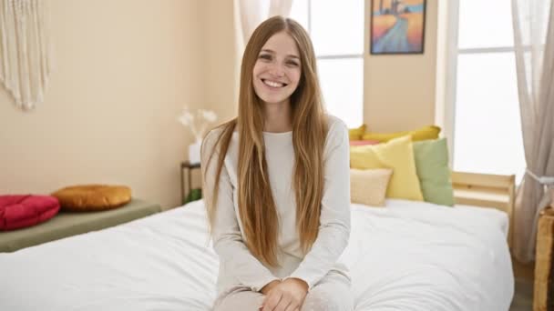 穿着睡衣快乐地坐在床上的年轻漂亮的金发女人 她那淡淡的微笑照亮了她舒适公寓里的房间 — 图库视频影像