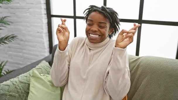 希望の若い黒人女性は 彼女の居心地の良いホームソファーに座って 運のために指を交差しています 目を閉じて ポジティブで陽気なジェスチャーを表現 — ストック動画