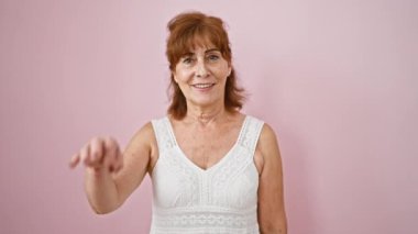 Neşeli orta yaşlı kadın neşeli bir el hareketi yapıyor, parmaklarından dürbün gibi bakıyor, izole edilmiş pembe arka planda mutluluk saçan gülen yüzü