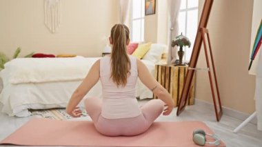 Bir kadın, nötr renk paleti olan rahat, aydınlık bir yatak odasında pembe bir paspasın üzerinde yoga egzersizi yapıyor..