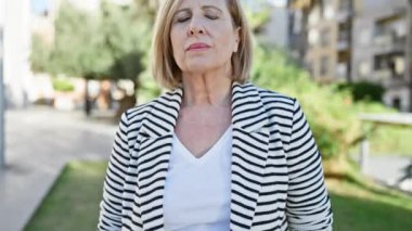 Olgun sarışın kadın, gözleri kapalı güneşli bir parkta huzur içinde duruyor.