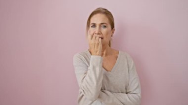 Gergin, orta yaşlı, endişeyle tırnak yiyen, pembe izole edilmiş arka planda duran, korku, endişe ve stres gösteren sarışın bir kadın.