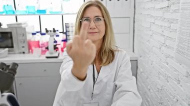 Mature, blonde scientist, in a lab, rudely flips the bird - a naughty 'fuck off' show of attitude. provocatively humorous, yet impolite middle-aged woman's expression.