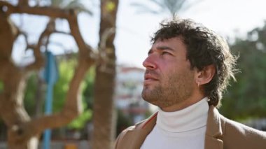 Sakallı mutlu bir İspanyol adam güneşli bir parkta sıcacık gülümsüyor, günlük şehir hayatını canlandırıyor..