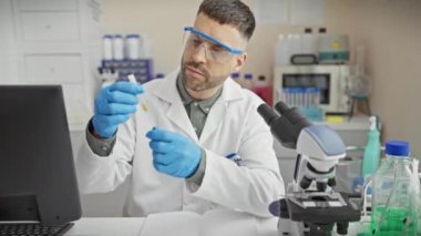 Laboratuvar önlüğü giymiş odaklanmış bir adam laboratuarda mikroskop ve bilgisayarla bir örneği inceliyor..