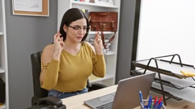 Gözlük takan kendine güvenen genç bir İspanyol kadın çalışma masasında heyecanlı bir şekilde çalışıyor, pozitifliğini ifade ediyor..