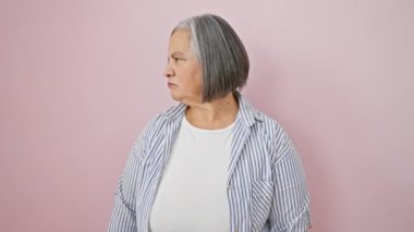 Rahat ama ağırbaşlı gri saçlı orta yaşlı kadın, sade gömlek giyiyor, pembe izole edilmiş arka planda ciddi yüz ifadesiyle kameraya ciddi bir bakış atıyor..