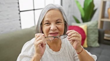 Evin içinde gülümseyen, oturma odası arka planında gözlük tutan son sınıf öğrencisi bir kadın.