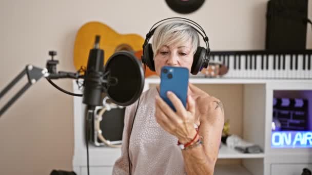 一个头发灰白 头戴耳机 手持智能手机 背景音乐工作室里演唱的成熟女人 — 图库视频影像
