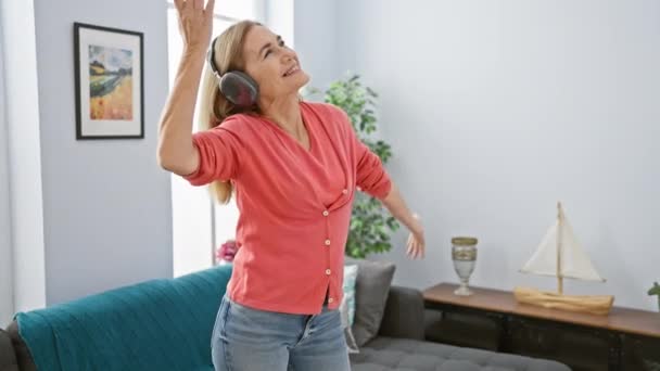 充满热情的成熟女人喜欢带着耳机在家里跳舞 在舒适的客厅里感到快乐 — 图库视频影像