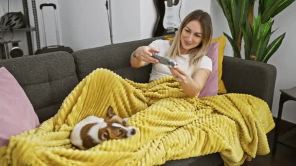 笑顔の若い女性は自宅のソファーで黄色の毛布でリラックスしながら彼女のかわいい犬の写真を撮ります — ストック動画