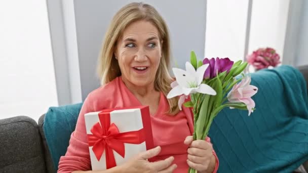 懐疑的な外観を持ち 自宅で贈り物や花束を保持し 彼女の顔に疑念とサルカズムのブレンドを披露する中年ブロンドの女性 — ストック動画