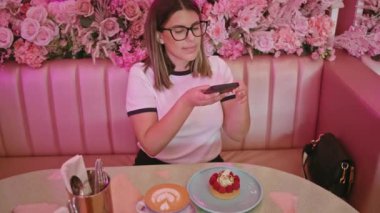 Çağdaş bir kadın akıllı telefonuna bakarken şık pembe bir kafede tatlı yemekten hoşlanır.