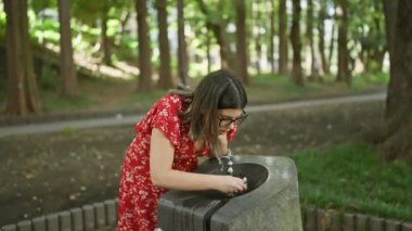 Parktaki çeşmeden serinletici bir içecekle susuzluğunu gideren göz kamaştırıcı İspanyol kadın, bardakları yazın sıçramasıyla parıldıyor! Güzel açık hava portresi, sağlıklı ve nemlendirici seçenekleri onaylıyor..