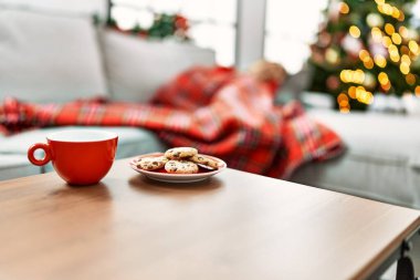 Kanepede uyuyan bir kadının olduğu rahat bir Noel sahnesi, kırmızı bir kupa, kurabiyeler, bokeh ışıkları, ve neşeli bir atmosfer..