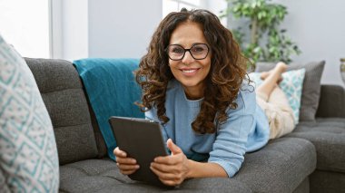 Kıvırcık saçlı, gülümseyen İspanyol kadın evdeki kanepede dinlenirken tablet kullanıyor..