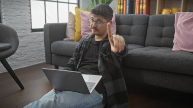 Young hispanic man with glasses and moustache using laptop in modern living room, showing thumbs down gesture. clipart