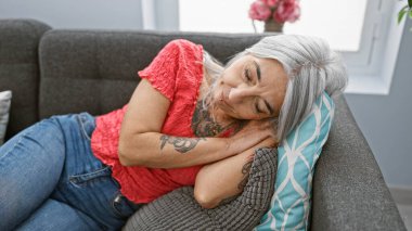 Relaxed, beautiful grey-haired middle age woman finds comfort in rest, lying down and sleeping on a cozy sofa indoors, home is her sanctuary where tiredness subsides clipart