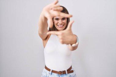 Beyaz arka planda duran İspanyol genç kadın gülümsüyor ve mutlu yüzlü eller ve parmaklarla çerçeve yapıyor. yaratıcılık ve fotoğrafçılık kavramı. 