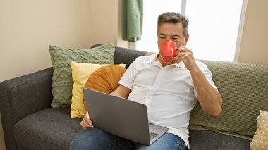 Rahat bir evde oturmuş laptopla çalışırken kahve içen, rahat bir telekomünikasyon sahnesi çizen olgun bir adam..