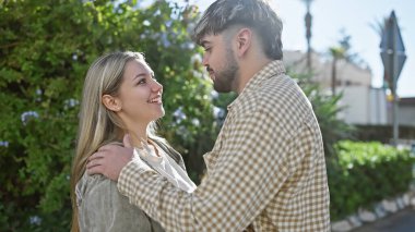 Gündelik giysiler içinde mutlu bir çift sevgiyle kucaklaşıyor, güneşli bir park yolunda bir anı paylaşıyor yeşilliklerle çevrili.