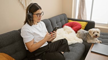 Kulaklıklı İspanyol kadın rahat bir oturma odasında köpeğin yanında akıllı telefon kullanıyor..