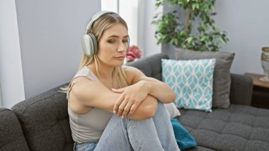 Ciddi yüzlü genç sarışın kadın evde oturmuş müziğin sesine dalmış, kendi oturma odasındaki kanepeye sarılmış bir şekilde derinden konsantre oluyor.