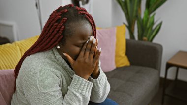 Üzüntülü bir Afrikalı Amerikalı kadın örgülü modern bir oturma odasında oturmuş elleriyle yüzünü kapatıyor..