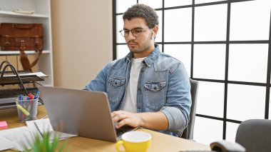 Gözlüklü ve kot pantolonlu bir adam iyi aydınlatılmış modern bir ofis odasında dizüstü bilgisayarda yazıyor, verimlilik ve konsantrasyon sergiliyor..