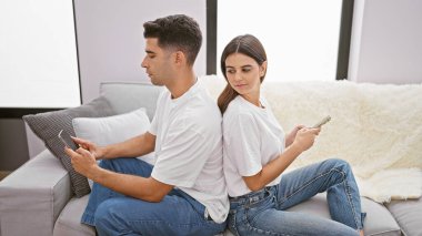 Bir erkek ve kadın, modern oturma odasındaki akıllı telefonları kullanarak kanepede sırt sırta otururlar. Bu da ilişkilerinde potansiyel bir kopukluk olduğunu gösterir..