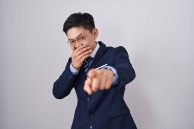 Genç Asyalı takım elbiseli ve kravatlı bir adam sana gülüyor, eli ağzında kamerayı işaret ediyor, utanç ifadesi. 