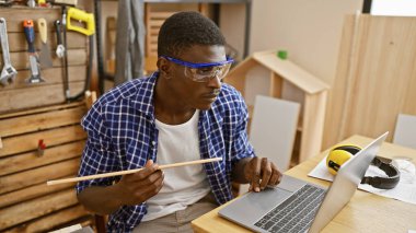 Güvenlik gözlüğü takan yakışıklı siyah adam marangozluk atölyesinde dizüstü bilgisayara odaklanıyor.