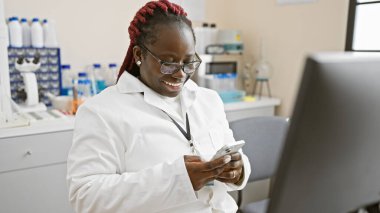 Örgülü, laboratuvar önlüğü giymiş, eczanede akıllı telefon kullanan gülümseyen Afrikalı Amerikalı kadın..