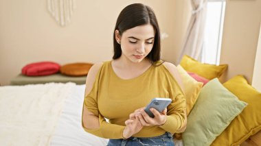 Çekici genç İspanyol kadın renkli yastıklı bir yatak odasında akıllı telefon kullanıyor..