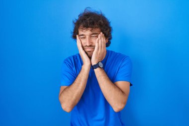 Hispanic young man standing over blue background tired hands covering face, depression and sadness, upset and irritated for problem 