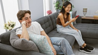 Modern bir oturma odasında akıllı telefon kullanan ilgisiz bir adam ve gülümseyen bir kadın ilişki dinamiklerini tasvir ediyorlar..