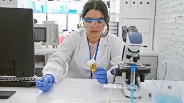 Odaklanmış bir İspanyol kadın bilim adamı, bir laboratuvar ortamında, gelişmiş ekipmanlarla çevrili bir örneği inceliyor..