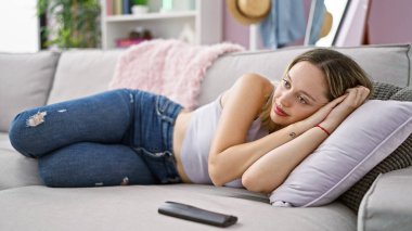 Kanepede uzanmış televizyon izleyen genç sarışın kadın evde gülümsüyor.