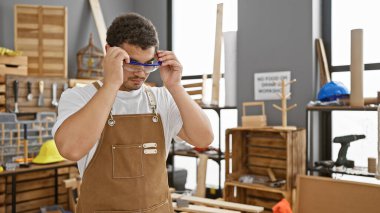 Ahşap mobilya ve aletlerle çevrili, iyi organize edilmiş bir marangozluk atölyesinde güvenlik gözlüğü ayarlayan genç bir adam..