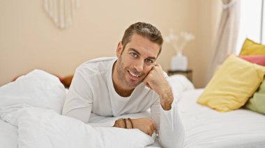 Beyaz gömlek giyen, sakallı neşeli bir adam, renkli yastıklarla parlak bir yatak odasında dinleniyor..