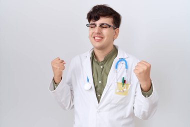 Doktor üniforması ve steteskop giyen genç, mutlu ve heyecanlı, kolları havada, gülümseyen ve başarı için bağıran bir jest yapıyor. kutlama konsepti. 